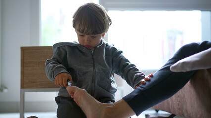 Child attempting to trim mother's toe nails, authentic domestic lifestyle scene of cute little boy using nail clipper - obrazy, fototapety, plakaty