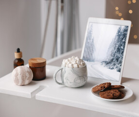 White wooden tray with tablet, cookies and spa products on bathtub in bathroom