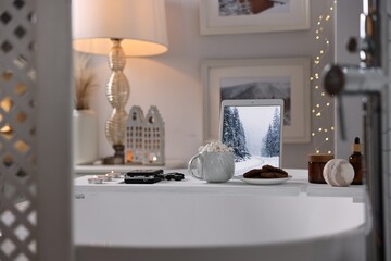 White wooden tray with tablet, spa products and burning candles on bathtub in bathroom