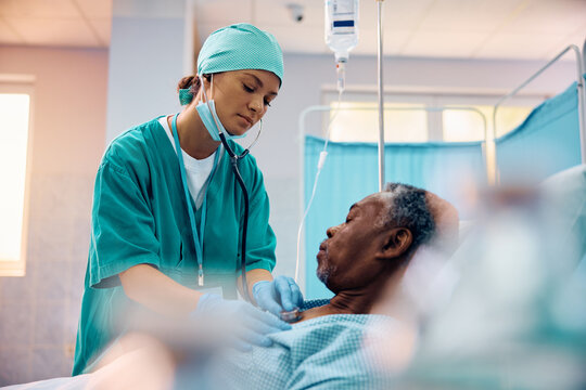 Female doctor using stethoscope while examining black senior patient in hospital ward.