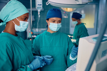 Female surgeons cooperating while looking at patient's vitals on monitor in operating room.