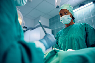 Black surgeon cooperating with her colleague during surgery in operating room. - obrazy, fototapety, plakaty