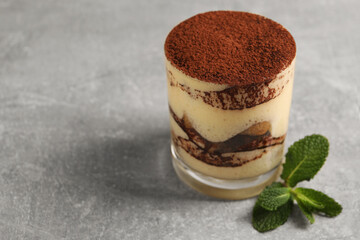 Delicious tiramisu in glass and mint on grey textured table, closeup. Space for text