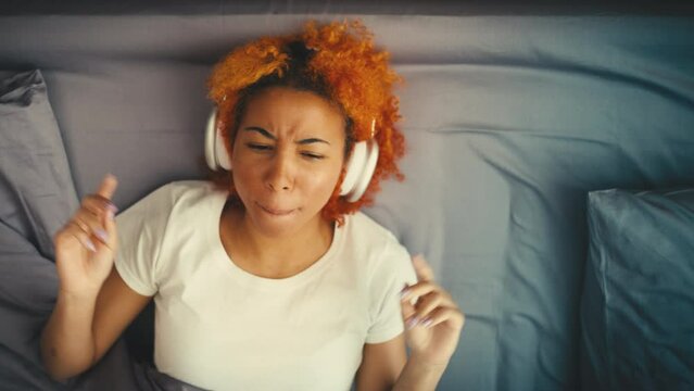 Curly-haired woman in headphones sings a song while lying in bed, musical hobby