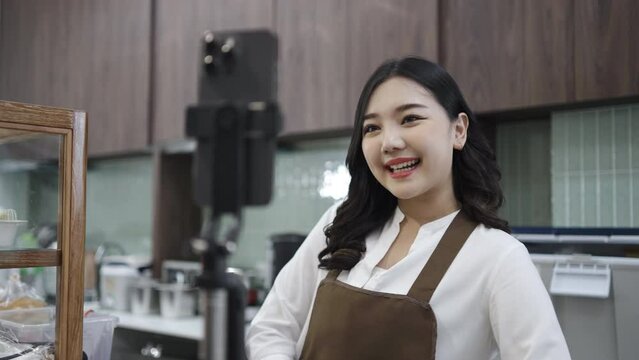 Young asian women is shop owner put on an apron show bread packages for sale while live streaming or recording video on social media in her cafe, coffee shop business concept.