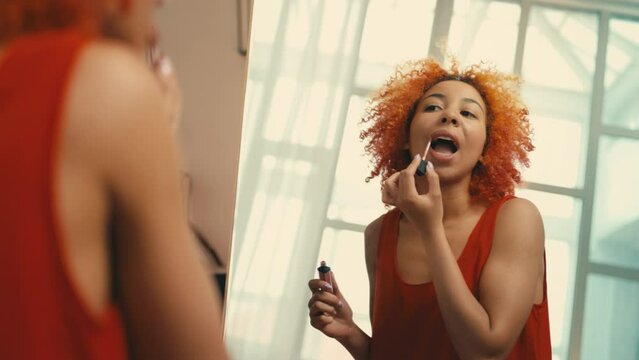 Happy woman applying lip gloss in front of mirror, getting ready for a party