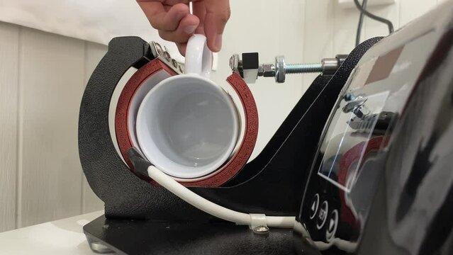 Male hand making a personalised white glass coffee mug with heat press printing machine.