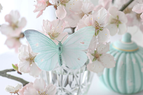 A soft blue butterfly perches delicately on cherry blossoms by a crystal vase. The image embodies the gentle rebirth of spring, ideal for tranquil decor themes