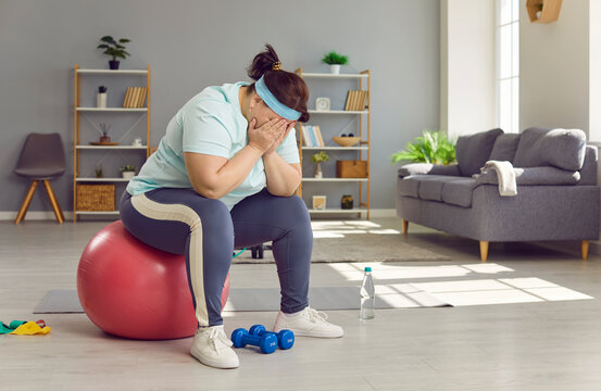 Fat woman can't lose weight despite her sports workouts. Sad, frustrated, exhausted, overweight, chubby, plump, young woman sitting on fitball in living room, hiding her face in hands, and crying