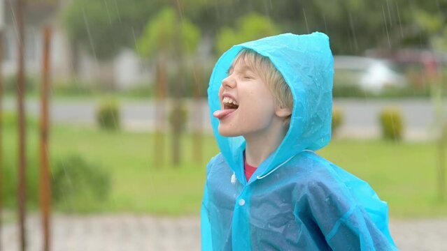 Portrait of a happy child in the pouring rain. A joyful boy in a blue raincoat catches water drops on his tongue in the pouring rain while walking. Children love to run and play in rainy weather