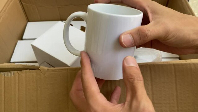 Male hands inspecting white glass coffee mug from many boxes of inventory.