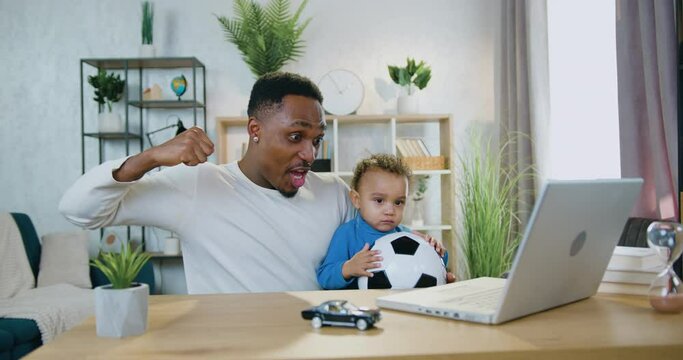 Likable African American man holding his calm baby boy on knees during watch football game on computer and shouting after scored goal