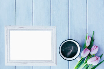 Blank empty picture frame with hot coffee and tulip flowers over blue rustic background with spring flowers. Table top view.
