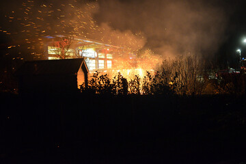 Riesiger Funkenflug aus dem qualmenden Feuer vor der Nachbar Feuerwache mit dicker Rauchwolke. Scharfer Vordergrund mit Gartenhaus und Hecke in Silhouette.