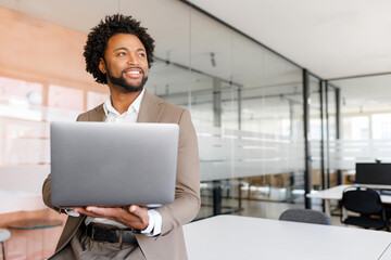 Poised African American businessman in a taupe suit holds his laptop and looks off into the...