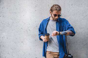 Portrait of cheerful young caucasian man holding coffee cup and checking time on watch while standing against grey background. Modern freelancer walking in the city - obrazy, fototapety, plakaty