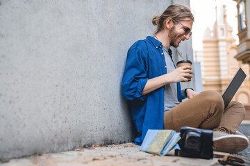 Side view shot of young stylish man work using laptop outdoors with coffee cup in hand. The concept of study, freelancing, work and travel. Modern student typing, doing projects online