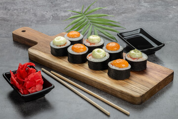 Bright maki rolls with white and orange sauce on a wooden board. Ginger, bamboo sticks and soy sauce on a gray tabletop.
