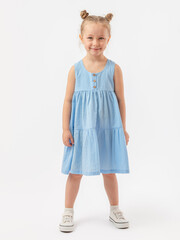Joy, fun. A beautiful 5-year-old girl with a fashionable hairstyle in a blue summer dress and sneakers stands on a white background and smiles. Legs apart.