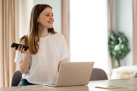 Cheerful Young Woman Multitasking With A Phone And Laptop, Embodying Flexibility And Efficiency.