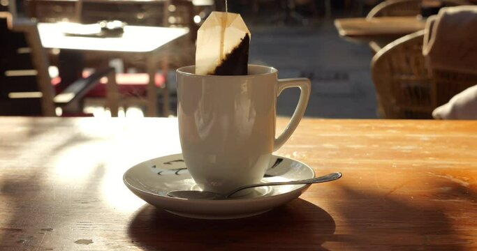 On sunlit morning at city cafe terrace, close-up of tea cup reveals teabag being slowly immersed and moved up and down in hot water. Aromatic drink steams as teabag is dunked, creating cozy ambiance