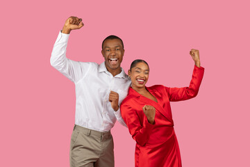 Joyful black couple celebrating with fists raised, dressed in white and red, pink background