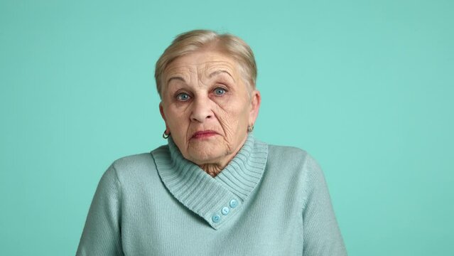 Puzzled old lady with short colored hair, neat eyebrows, light make up wearing blue sweater standing in studio shrugging shoulders with misunderstanding. High quality 4k footage