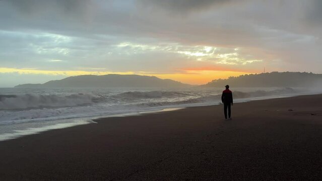 A man walks along a sandy beach along a stormy sea against the backdrop of a beautiful golden sunset.