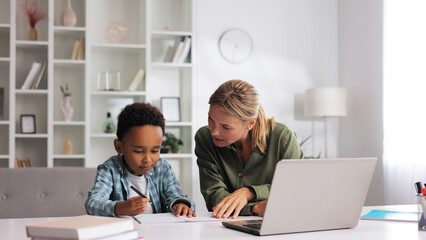Blonde tutor teaching little African American preschooler to write at home. Help with early childhood education in an online school. Happy boy is learning to write.