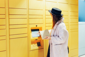 Fashion woman with box using modern postal automatic mail terminal with self service device for pickup or refund an order. Electronic locker for storing parcels. Online shopping. Selective focus.