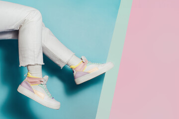 Close up female legs in white jeans and retro style high-top multicolor sport sneakers shoes on multicolor blue and pink background. Pastel candy colors, vintage retro style of 80s - 90s vibes. - obrazy, fototapety, plakaty