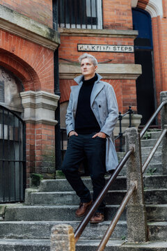 Handsome Male Model With Gray Hair In A Black Roll Neck Jacket And Trousers, Checked Coat Walking On The Old City Street. Cold Season Fashion Stylish Outfit Trend. Old Money Aesthetic. Soft Focus