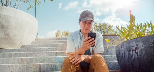 A young man in casual stylish clothes sits on the steps of a modern park on a sunny day and holds a...