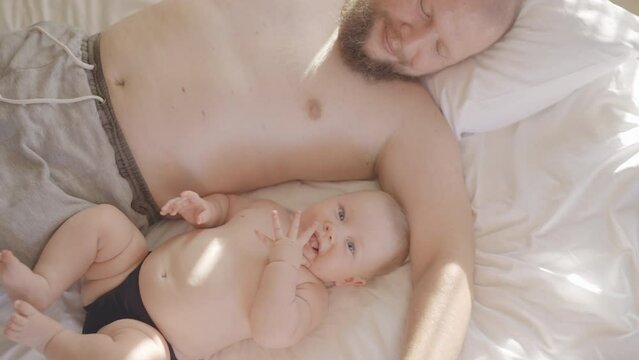Young father lying in bed caressing little baby boy. Daddy kiss play with Newborn child. Family, parenthood, childhood. Touching tender moment of love, soft touch baby skin. Authentic shot loving dad