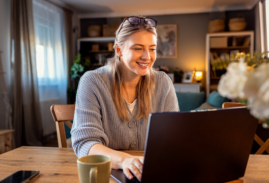 Smiling young woman working at home using laptop while sitting in her living room.