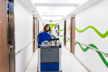 A warm-hearted nurse behind a coffee service cart in a hospital corridor, with a doctor in the soft-focused background. © Koldo_Studio