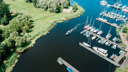 Aerial view of a peaceful marina with various sailboats docked along the pier, surrounded by lush greenery on a bright sunny day