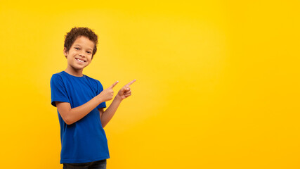 Latin boy with bright smile pointing at copy space against yellow backdrop