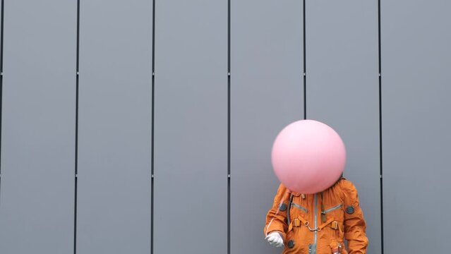 Happy astronaut wearing space suit with helmet hiding head under helium balloon outdoors. Bizarre concept