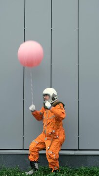 Astronaut wearing space suit with helmet dancing with inflatable balloon in hand outdoors. Surreal concept