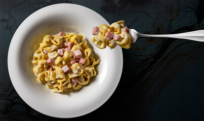 White soup plate and fork with tortellini with cream and ham on black background
