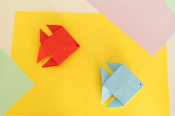 Two origami fish, one red and one blue, positioned playfully on a vivid yellow background with soft...
