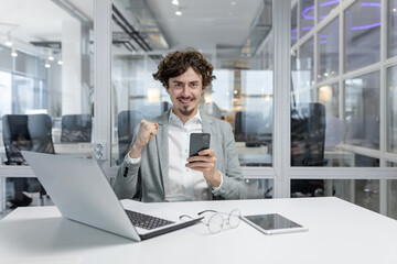 Enthusiastic young adult working at his office desk, showing victory gesture with a smartphone in...