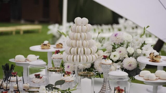 elegant dessert table at an outdoor event, with a clear focus on a macaron tower surrounded by various desserts, candles, and a lush floral arrangement. The white and green color palette suggests 