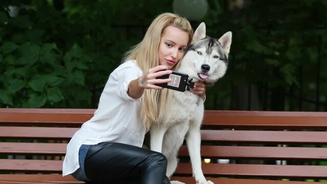 Young woman with dog Husky doing selfie on bench in park.