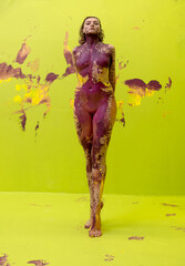 Young artistic sexy proud nude woman in yellow and magenta paint, standing in front of the light...