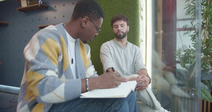 Handsome, black, male employee drawing his white colleague in a notebook during break at work while sitting on a sofa at the threshold in the office. Talented employee drawing portrait of colleague.