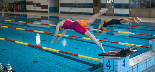 Two professional female swimmers taking track start position on the platform gracefully jumping and diving in a swimming pool. Sport competition concept.