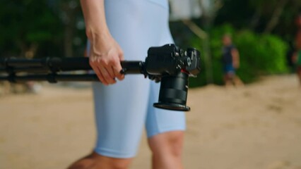 Woman explores beach, searches ideal shot with camera on tripod. Vibrant scenery, traveler captures visuals for blog, engaging viewers. Unique angle, creative content creation on sandy shore.