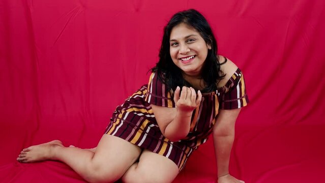 Smiling woman sitting against a red background wearing a striped dress looking down with a joyful expression